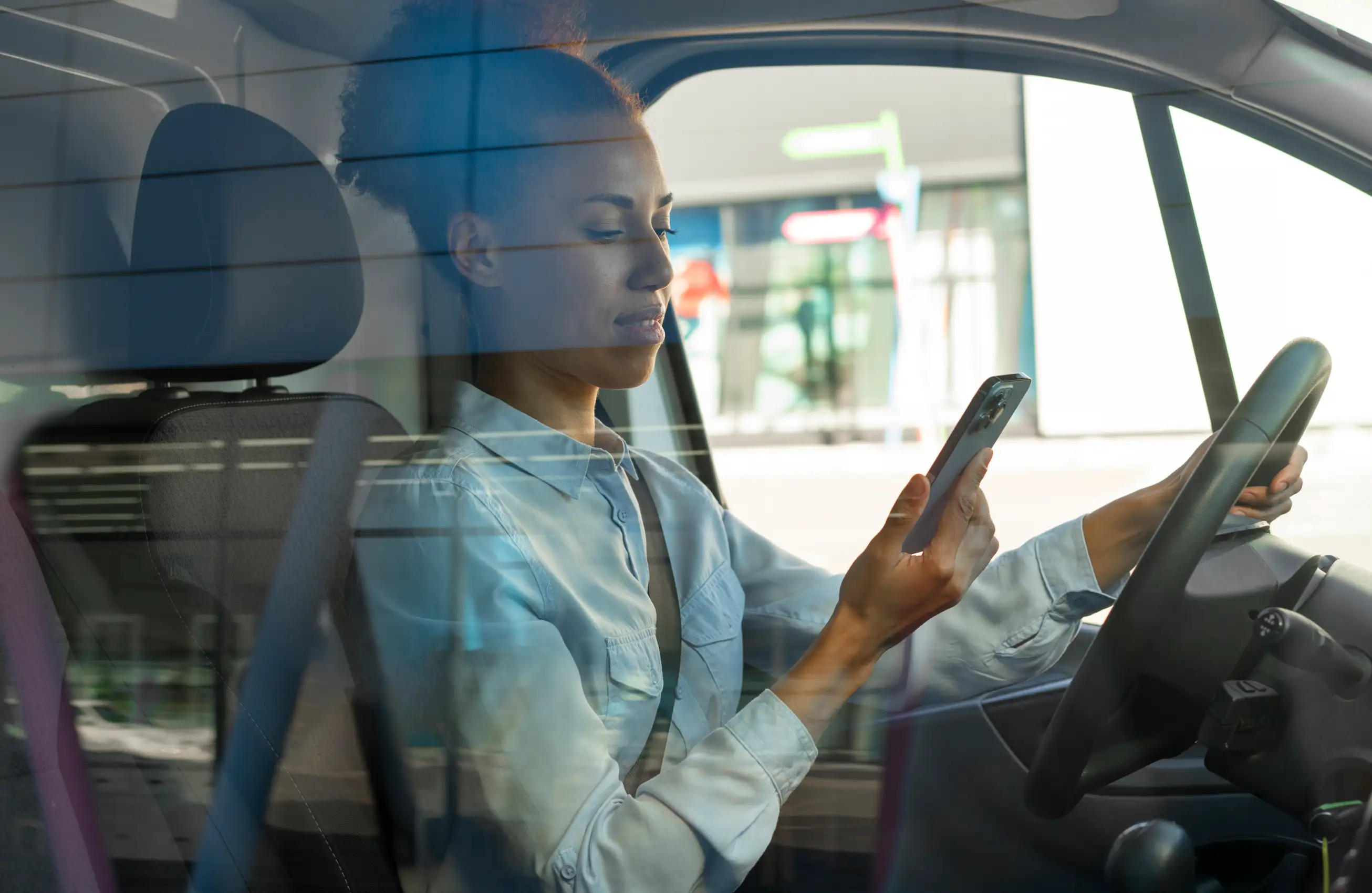 Mulher sentada ao volante de um carro segurando um smartphone com a mão direita enquanto dirige com a esquerda, vista pelo vidro do veículo.