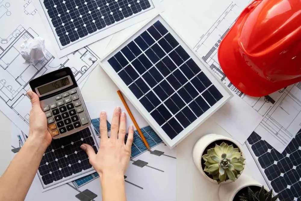 Mesa com projetos de painéis solares, uma calculadora, uma planta suculenta e um capacete vermelho, simbolizando a transferência de créditos de energia solar.