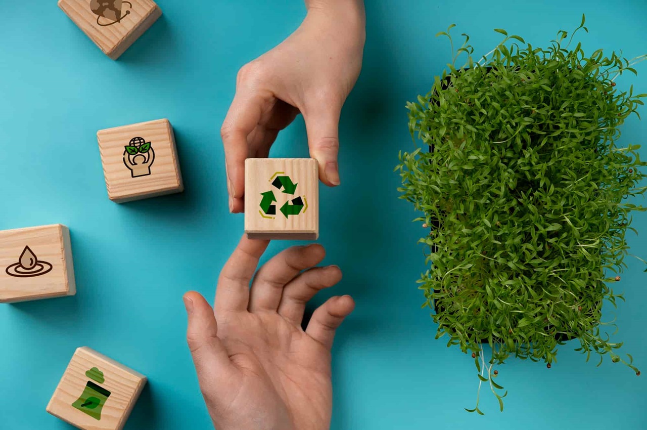 Mãos trocando blocos com símbolos de reciclagem e sustentabilidade, ao lado de uma planta.
