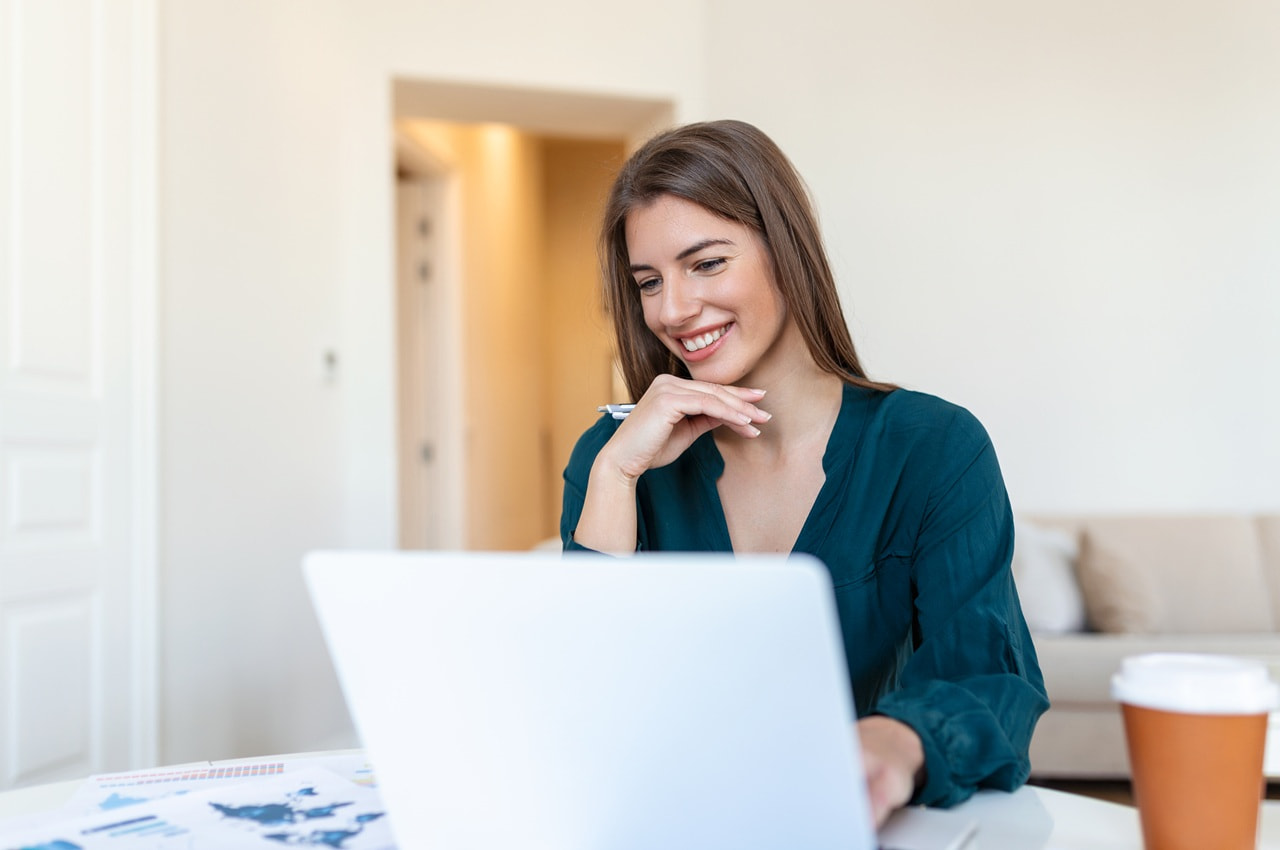 Mulher com uma blusa verde-azulado sorrindo enquanto usa um notebook em uma mesa clara.