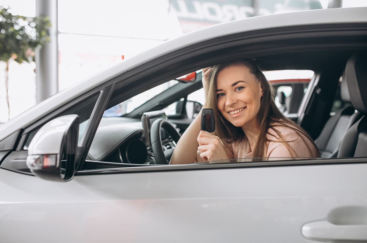 Mulher dentro de um carro branco, sorrindo e mostrando a chave do veículo leve.