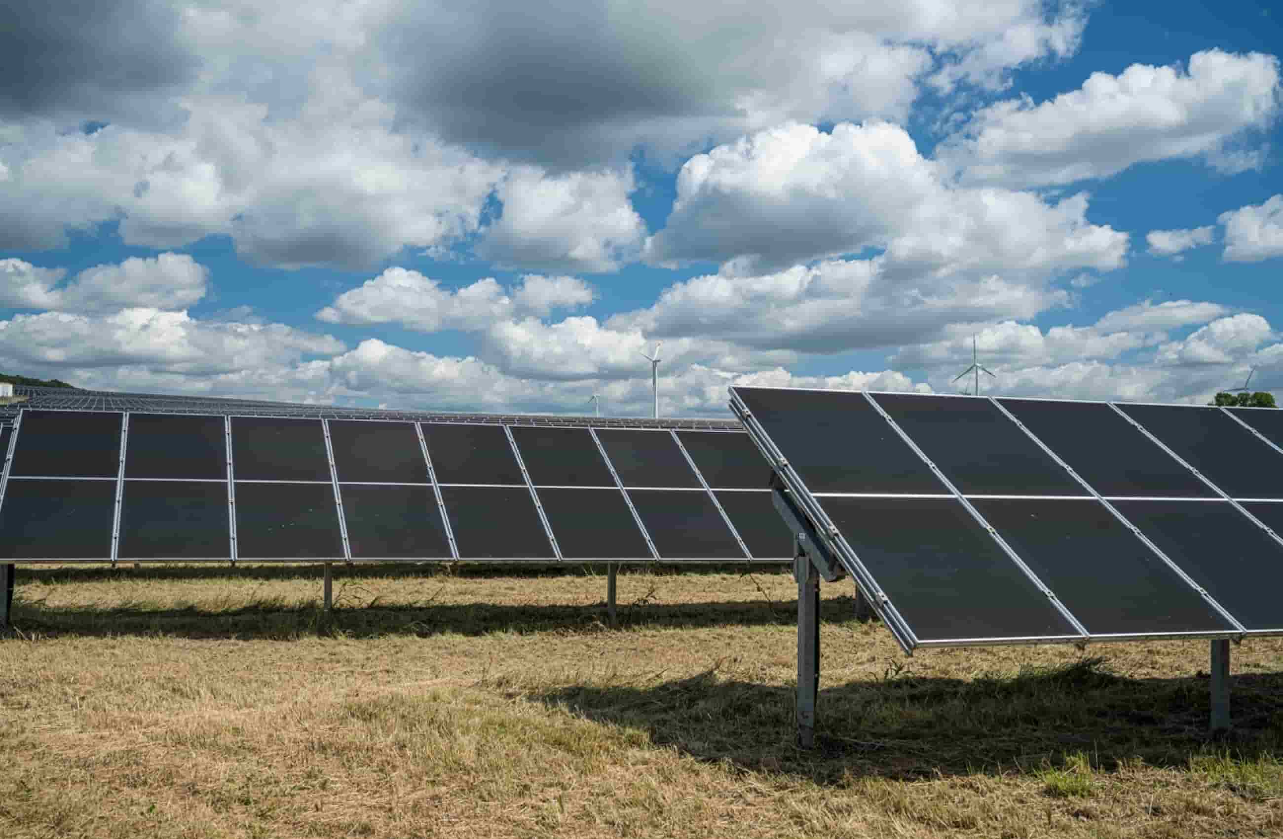 Duas placas solares grandes instaladas em um campo de grama. A cima delas tem algumas nuvens no céu azul.
