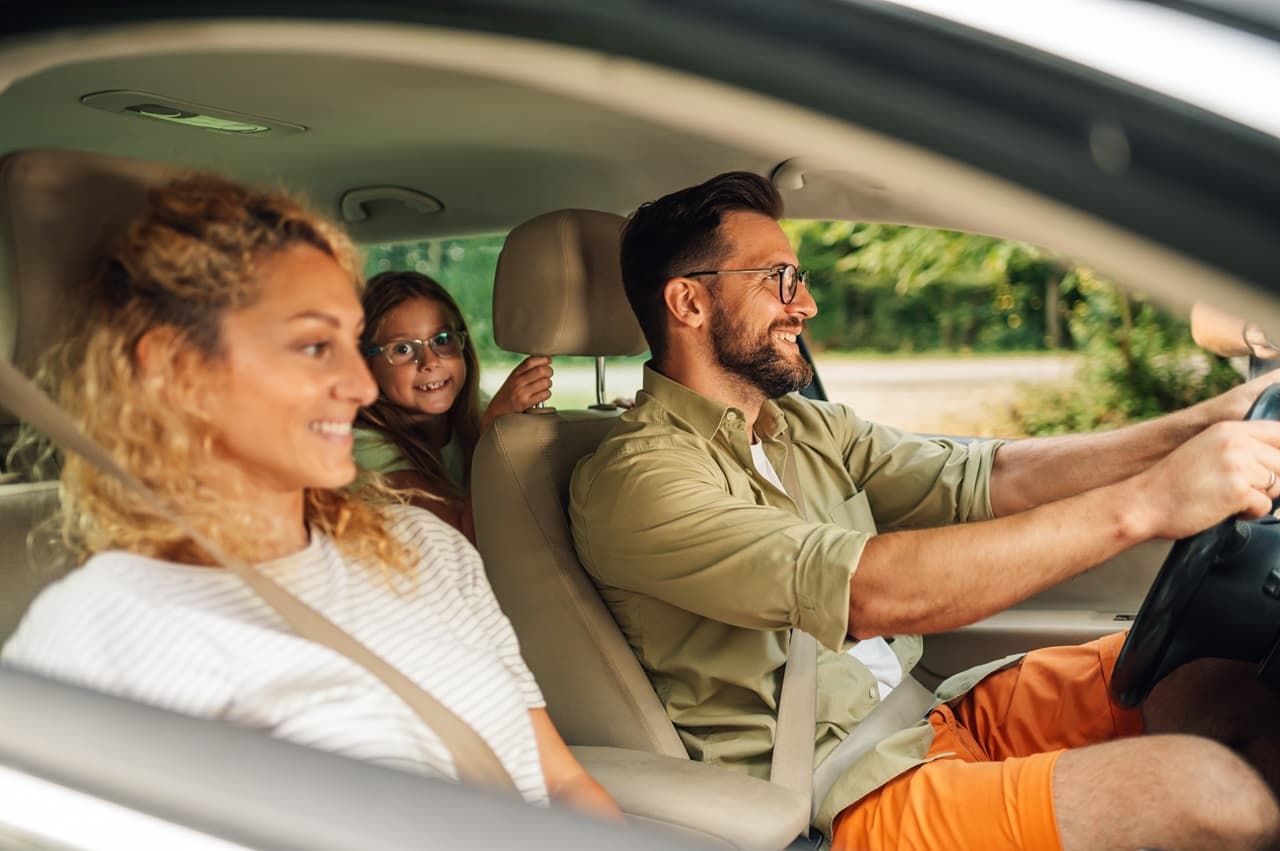 Homem dirigindo carro, com mulher ao seu lado no banco de passageiro e menina no banco de trás. Todos estão sorrindo.