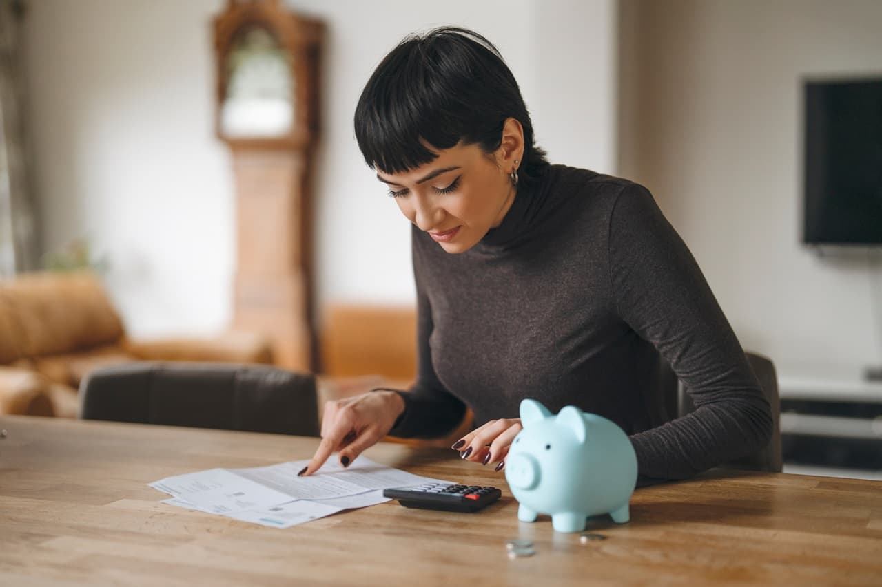 mulher sentada de frente para uma mesa com um cofrinho em cima, analisando contas com uma calculadora