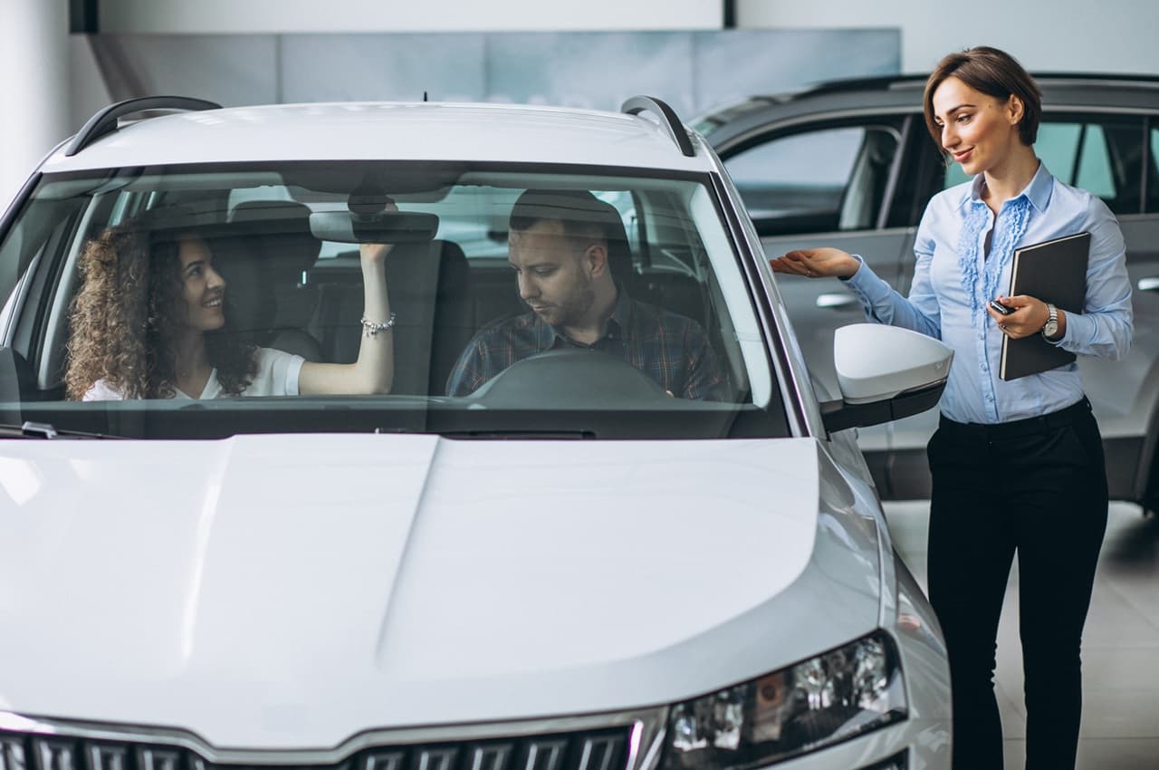 Vendedora apresentando o valor venal de um carro branco para um casal, que está no interior do veículo na concessionária.