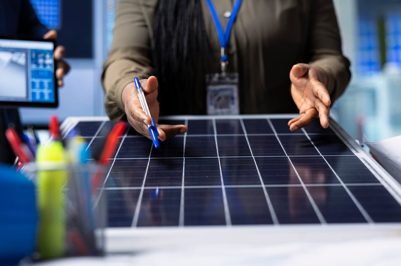 Mãos de uma mulher segurando uma caneta e gesticulando sobre a superfície de um painel solar fotovoltaico.