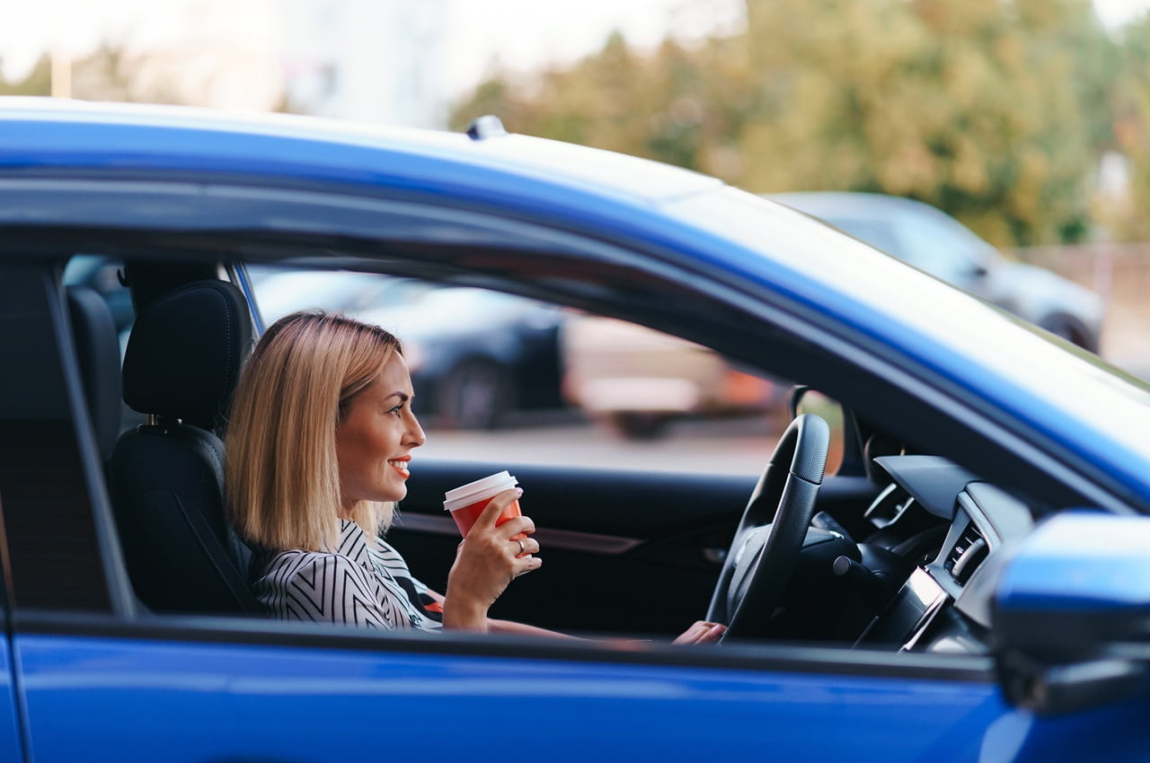 Mulher loira sentada ao volante de um carro azul. Ela está segurando um copo de café e sorrindo.