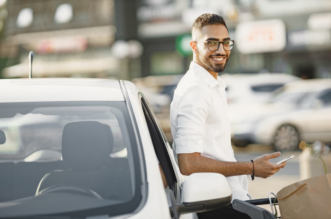 Homem de óculos e camisa branca encostado na lateral de um carro branco, enquanto segura um celular com uma das mãos.