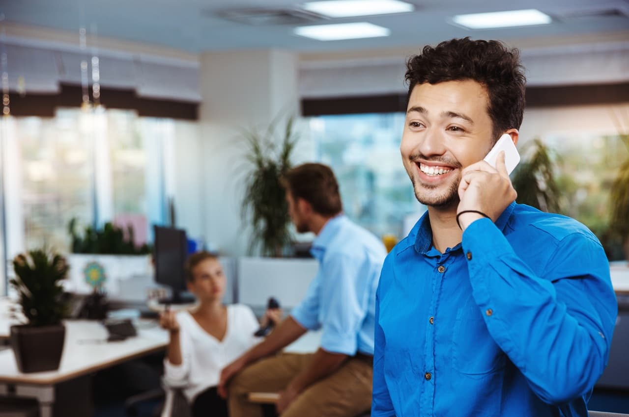 Homem jovem de camisa azul sorrindo enquanto fala ao celular em um escritório, com colegas conversando ao fundo.