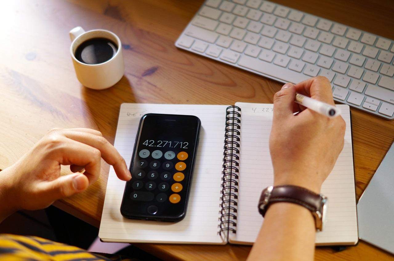 A imagem mostra a visão das mãos de uma pessoa, escrevendo em uma agenda e usando a calculadora em seu celular. Além disso, também existe uma xícara de café e um teclado de computador.