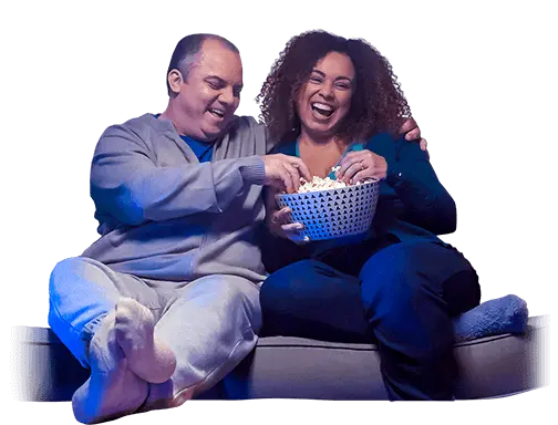 Um homem e uma mulher sentados no sofá, rindo e comendo pipoca, faça como eles e organize suas finanças com um crédito rápido aqui na BV