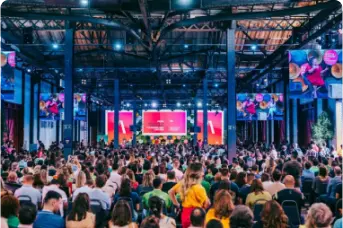 Imagem do logotipo do evento Web Summit Rio em branco e laranja. Ao fundo temos o local do evento, Riocentro.